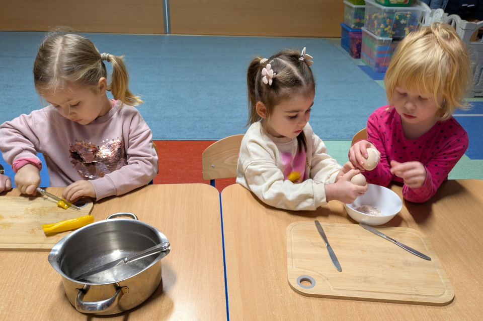 Trzy dziewczynki siedzą przy stole - dwie obierają jajka, jedna kroi paprykę