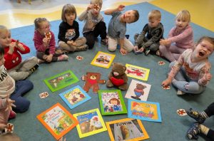 Na dywanie rozłożone książki dla dzieci, na samym środku siedzi pluszowy miś. Wokół książek siedzą dzieci i trzymają w rękach papierowe misie