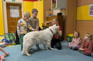 Biały owczarek szwajcarski stoi bokiem, dwója chłopców głaska go, trenerka psów patrzy na dzieci