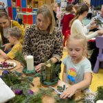 Dziewczynka na mamą robi stroik świąteczny. Na drugim planie inni rodzice z dzieći