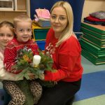 Dziewczynka trzyma chłopca na kolanach, mama przykuca obok. Prezentują stroik świąteczny